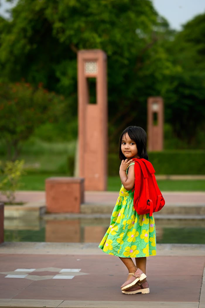 A young girl in a summer dress and red backpack enjoys a day at the park.
